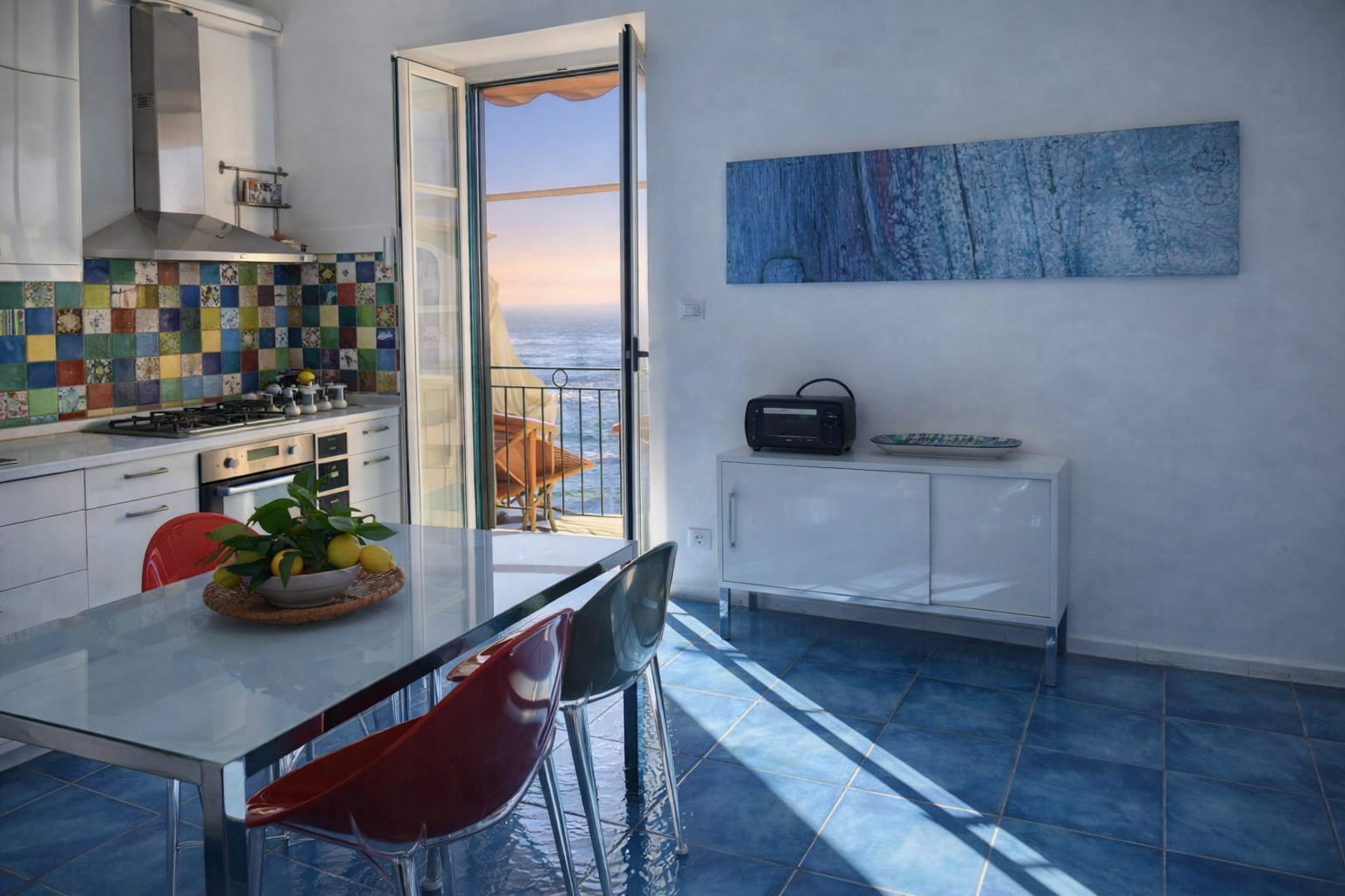 Cucina di AMALFI Sea House con piastrelle colorate tipiche amalfitane, porta aperta sul balcone con vista mare al tramonto, limoni sul tavolo e pavimento in maiolica blu