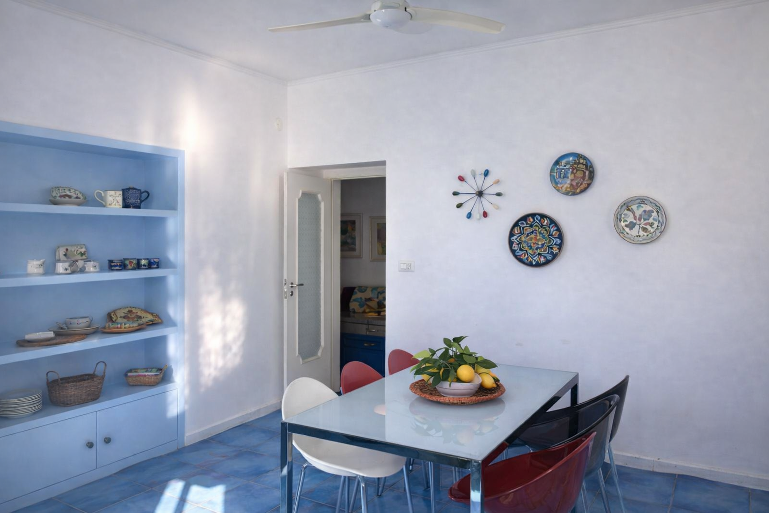Sala da pranzo mediterranea di AMALFI Sea House con tavolo in vetro, sedie design, piatti in ceramica dipinta su parete e libreria in blu con collezione di ceramiche locali amalfitane
